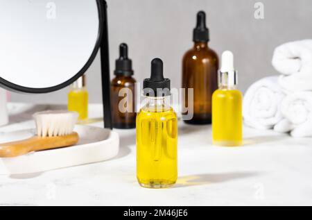Olii, spazzola da bagno, asciugamano e specchio sul tavolo cosmetico in bagno. Set di cosmetici organici in fiale di vetro con contagocce per la cura della pelle e dei capelli Foto Stock