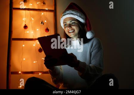 Donna che indossa il cappello Santa usando un tablet digitale a casa nel tablet della notte di natale illuminando il volto della donna Foto Stock