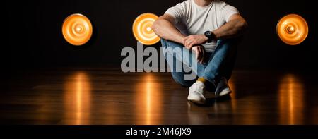 Uomo alla moda e irriconoscibile in jeans e una T-shirt bianca si siede sul pavimento in una stanza buia. Foto Stock