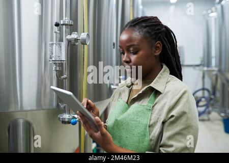 Giovane donna nera che si riferisce al computer tablet durante il controllo del processo di fermentazione Foto Stock
