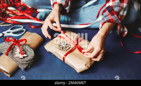 Le mani della donna che legano l'arco sul regalo di Natale imballato in carta marrone del mestiere. Foto Stock