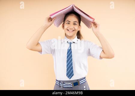 Happy studentessa indiana scuola che indossa uniforme scuola tenendo libri in piedi in testa isolato su sfondo beige, Studio shot, concetto di istruzione. Foto Stock