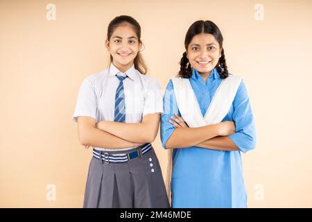 Felice rurale e moderna studentesse indiane che indossano uniforme scuola in piedi insieme braccia incrociate isolate su sfondo beige, Closeup, Studio Foto Stock