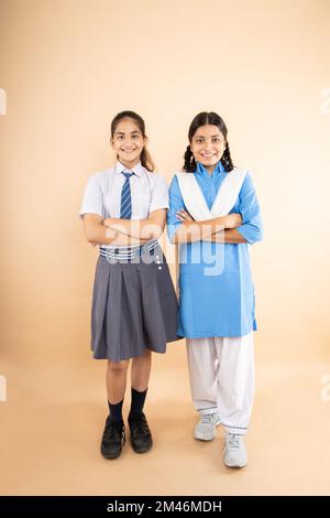 Happy Rural e moderna studentessa indiana con uniforme scuola in piedi mani piegate insieme isolato su sfondo beige, lunghezza completa, S Foto Stock