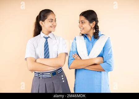 Buone studentesse indiane rurali e moderne che indossano l'uniforme della scuola in piedi insieme le braccia incrociate che si guardano isolate sopra il dorso beige Foto Stock