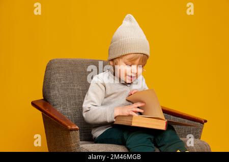 Ritratto di ragazzino, bambino in posa, seduto su sedia e libro di lettura su sfondo giallo studio. Fiabe Foto Stock