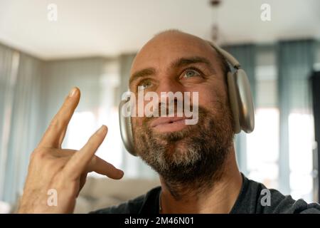 Uomo maturo che guarda con un sorriso e un rock sign mentre ascolta la musica preferita con le cuffie wireless a casa Foto Stock
