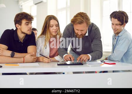 Un insegnante di sesso maschile in classe aiuta un gruppo di studenti delle scuole superiori a lavorare su un tablet digitale. Foto Stock