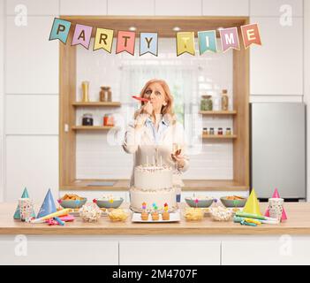 Casual donna matura soffiare un corno partito dietro un bancone con torta e decorazioni all'interno di una cucina Foto Stock