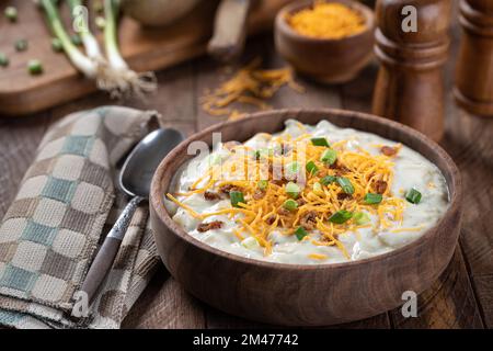 Zuppa di patate cremosa con pezzi di pancetta, formaggio cheddar grattugiato e cipolle verdi tritate in un recipiente di legno Foto Stock