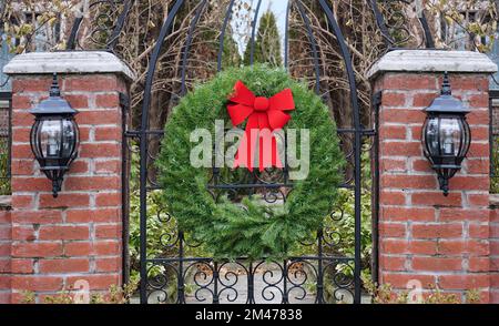 Cancello da giardino con colonne in mattoni decorate con corona di Natale con rami di pino e arco rosso Foto Stock