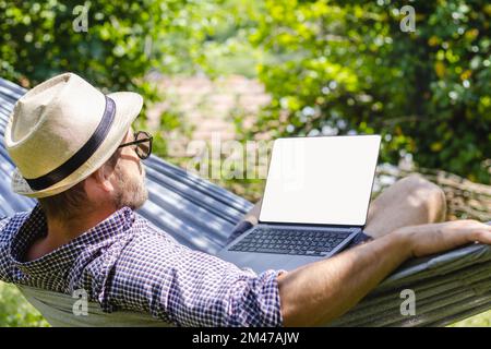 Uomo in cappello sdraiato in amaca in giardino e utilizzando il suo computer portatile. Foto Stock