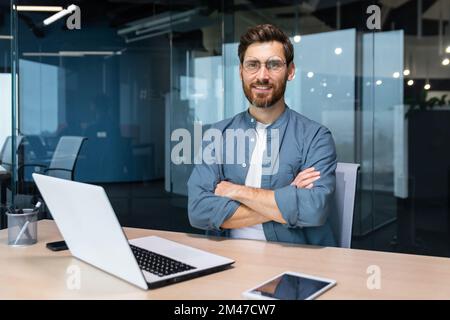 Ritratto di uomo d'affari maturo con barba, uomo in camicia sorridente e guardando la fotocamera seduto alla scrivania con braccia incrociate all'interno di un moderno edificio per uffici, utilizzando un tablet e un computer portatile al lavoro. Foto Stock