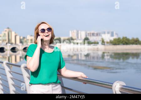 Una donna sorridente in occhiali da sole, parlando al telefono con gli amici gesturing, facendo data sulla riva del fiume in città. Ragazza sta camminando lungo il fiume della città Foto Stock