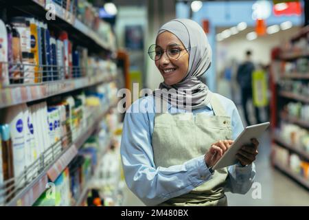 Una donna controlla i prodotti e le merci in un supermercato, una donna musulmana in un hijab usa un tablet vicino a uno scaffale con le merci, un lavoratore in un grembiule sorride al lavoro. Foto Stock