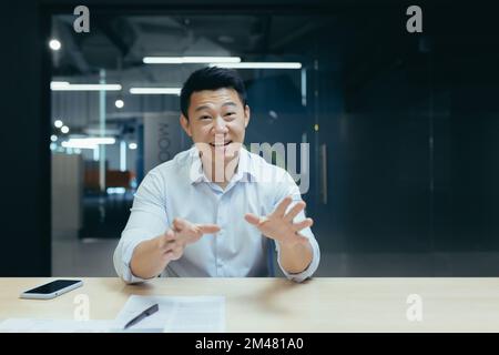Giovane asiatico sorridente seduto in ufficio alla scrivania e parlando con la fotocamera. Conduce riunioni online, consulti, corsi di formazione aziendale. Foto Stock