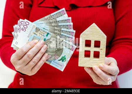 Il concetto del costo di possedere un appartamento in Polonia, Una casa di legno tenuta in mano e un pacchetto di 500 banconote zloty, il concetto di hou alto Foto Stock