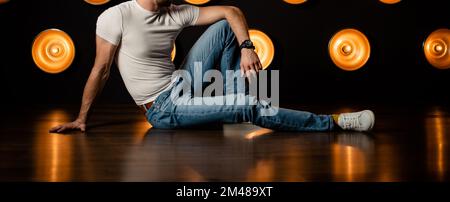 Uomo alla moda e irriconoscibile in jeans e una T-shirt bianca si siede sul pavimento in una stanza buia. Foto Stock