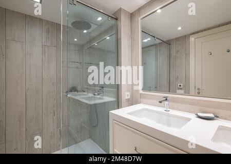 Bagno spazioso con illuminazione artificiale luminosa, grande specchio che riflette la porta anteriore e coppia di lavelli bianco neve incorporati in un armadio beige chiaro Foto Stock