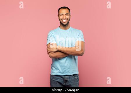 Uomo sicuro. Ritratto di felice afro americano ragazzo in piedi con braccia piegate e sorridente, posando su sfondo rosa Foto Stock