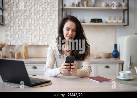 Giovane donna caucasica che controlla la posta sullo smartphone e che lavora su un computer portatile in interni di cucina minimalista, spazio libero Foto Stock