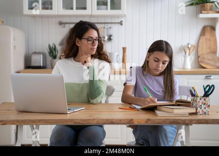 Madre che guarda la figlia facendo i compiti mentre lavora a distanza accanto, seduti insieme a un tavolo Foto Stock