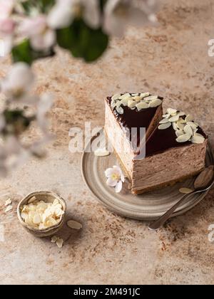 Due pezzi di cheesecake al cioccolato con petali di mandorla su piastra in ceramica su fondo marrone testurizzato. Fiori primaverili in sfocatura. Spazio di testo, menu. Vertica Foto Stock