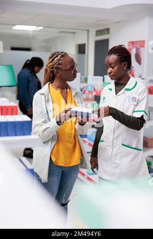 African american farmacista raccomandare farmaci donna in farmacia. Cliente e consulente di farmacia in piedi in corsia negozio di farmacia, cliente acquistare farmaci Foto Stock
