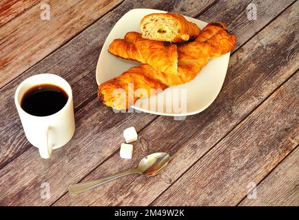 Un piatto con due focaccine di pasta sfoglia a spirale, una tazza di caffè caldo con un cucchiaio accanto e due cubetti di zucchero su un tavolo di legno. Primo piano. Foto Stock