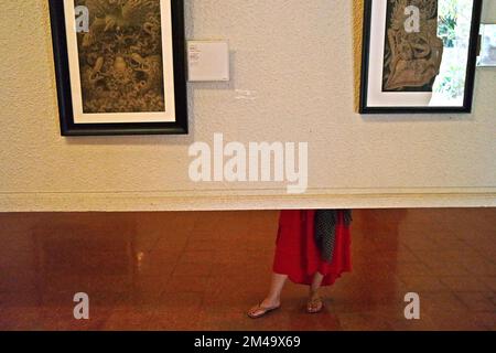 Una donna visitata viene fotografata da dietro una parete di esposizione portatile mentre si sta levando in piedi per prestare attenzione ai dipinti al Museo Puri Lukisan (museo del palazzo dei dipinti) a Ubud, Gianyar, Bali, Indonesia. Foto Stock