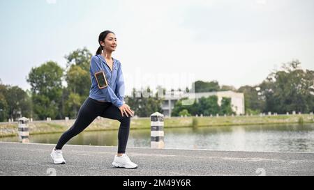 Sana e bella runner asiatica in abbigliamento sportivo allungando le gambe e le ginocchia, scaldandosi o raffreddando, preparandosi a correre in città pa Foto Stock