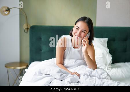 Bella giovane donna coreana che chiama, avendo una telefonata a letto, parlando con un amico mentre si soggiorna in camera da letto Foto Stock