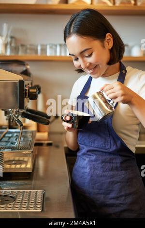 Primo piano di carina barista asiatica ragazza fare cappuccino, fare latte art in tazza con latte al vapore, in piedi in caffetteria dietro il bancone Foto Stock