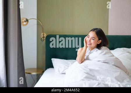 Bella ragazza asiatica sonnolenta si sveglia nel suo letto con comode coperte bianche, guardando fuori finestra con un sorriso soddisfatto. Concetto di stile di vita Foto Stock