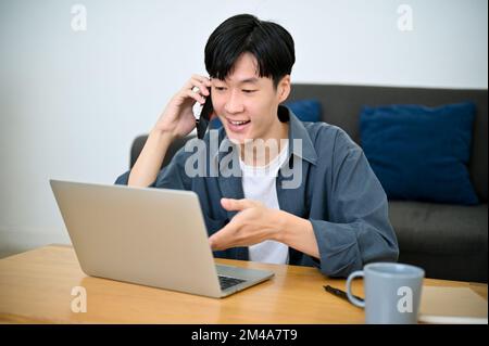 Felice giovane asiatico libero professionista che parla al telefono con il suo cliente mentre guarda alcuni dettagli sullo schermo del notebook. concetto di lavoro da casa Foto Stock