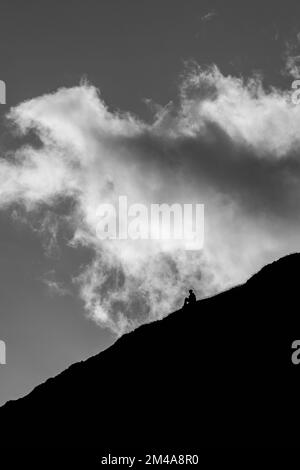 Una scala di grigi verticale di una persona seduta sul pendio di una montagna contro un cielo drammatico Foto Stock