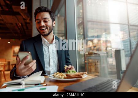 Un uomo d'affari indiano sorridente che indossa un costume usa il telefono durante il pranzo nel bar. Sfondo sfocato Foto Stock