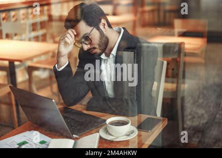 Un uomo d'affari indiano concentrato che lavora su un computer portatile in un accogliente bar vicino alla finestra Foto Stock