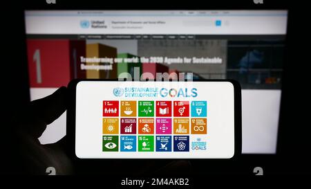 Persona che tiene in mano uno smartphone con il logo degli obiettivi di sviluppo sostenibile delle Nazioni Unite (SDG) sullo schermo di fronte al sito Web. Messa a fuoco sul display del telefono. Foto Stock