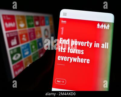 Persona che tiene in mano uno smartphone con la pagina Web degli obiettivi di sviluppo sostenibile delle Nazioni Unite (SDG) sullo schermo davanti al logo. Messa a fuoco sul centro del display. Foto Stock