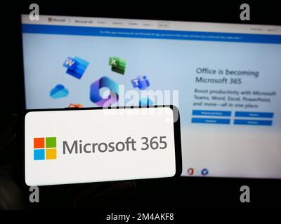 Persona che tiene lo smartphone con il logo del software di produttività Microsoft 365 sullo schermo davanti al sito Web. Messa a fuoco sul display del telefono. Foto Stock