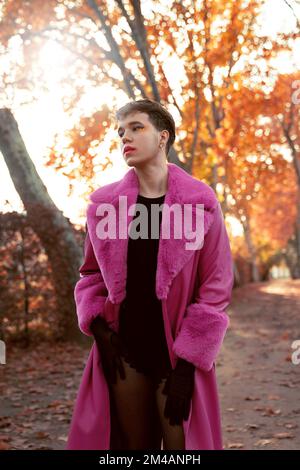 Auto assicurato giovane sesso maschio fluido con capelli corti e trucco in pelle fucsia brillante e pelliccia cappotto in piedi vicino agli alberi autunnali e guardando via sopra Foto Stock