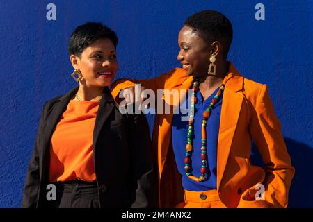 Donna afro-americana positiva in tuta arancione con capelli corti e orecchini in piedi vicino alla parete blu e mettendo mano sulla spalla della girlfrie ispanica Foto Stock