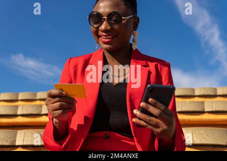 Donna afro-americana sorridente adulto positiva in abiti eleganti e occhiali da sole con carta di credito che effettua l'acquisto online via smartphone mentre si siede o Foto Stock