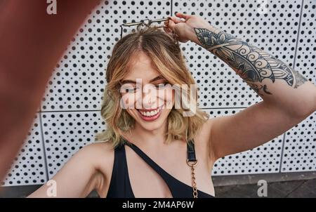 L'angolo alto di auto assicurato giovane allegro tatuato femminile millenario con capelli biondi sorridente brillante e regolare occhiali da sole mentre si prende selfie su Foto Stock