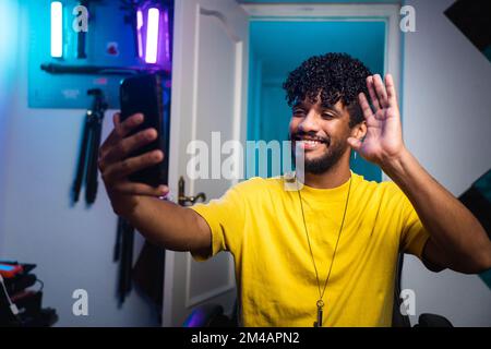 Allegro giovane uomo ispanico con capelli ricci sorridendo e ondeggiante mano mentre si siede alla scrivania dietro smartphone e riprese video per blog a casa Foto Stock