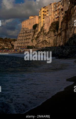 Tarda sera Agosto luce del sole e ombre mentre il mar Tirreno oscura lambisce una spiaggia ancora affollata di turisti sotto la spettacolare cittadina e località turistica sulla scogliera di Tropea, sulla Costa degli dei nella Calabria occidentale, nel sud Italia. Foto Stock