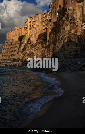 In una calda tarda serata ad agosto, i turisti iniziano a lasciare la spiaggia di Convento a Tropea, nella Calabria occidentale, nel sud dell'Italia, mentre le ombre che scuriscono la superficie del Mar Tirreno scivolano sulle sabbie e le nuvole scure si raccolgono sulla storica città arroccata sopra di loro su scogliere di arenaria. Foto Stock