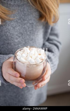 una ragazza in un maglione grigio tiene un bicchiere di cacao e marshmallows al mattino presto sullo sfondo della cucina da vicino. Foto Stock