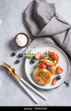 Tradizionale piatto russo per la colazione sciroppi. Frittelle al formaggio casolare con panna acida e frutti di bosco freschi su sfondo blu con stoviglie e pisolino Foto Stock
